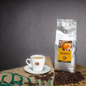White coffee cup on a saucer beside a tall silver Herbst coffee bag, with roasted beans scattered on a burlap surface.